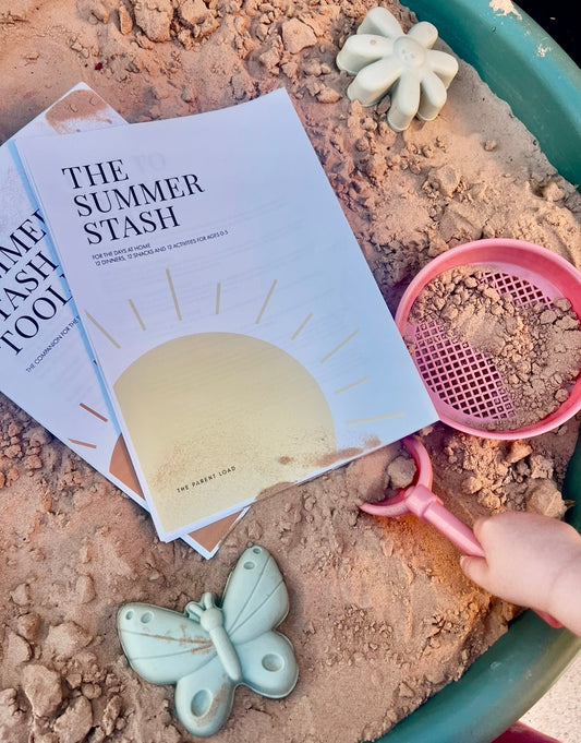 A styled flat-lay of a magazine or book titled "THE SUMMER STASH" on sand, alongside a pink scoop, a butterfly-shaped object, a flower-shaped object, and a teal container, with a hand holding the scoop.
